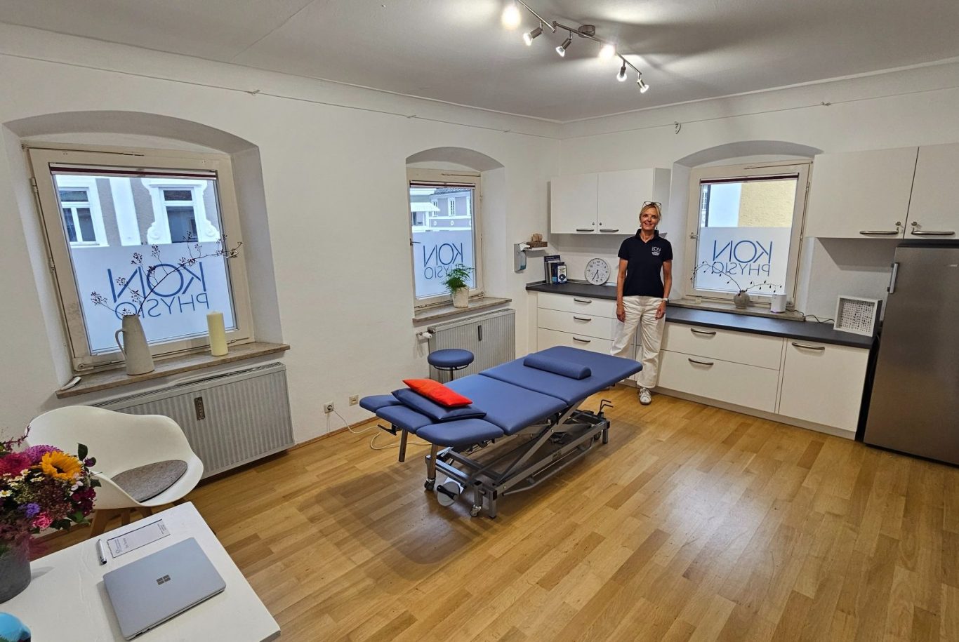 Physiotherapie Prien am Chiemsee Kathrin Ortinger Naujeck Lichtdurchfluteter, freundlicher Physiotherapieraum mit drei Fenstern, Parkett, moderne Behandlungsliege und Kathrin Ortinger Naujeck
