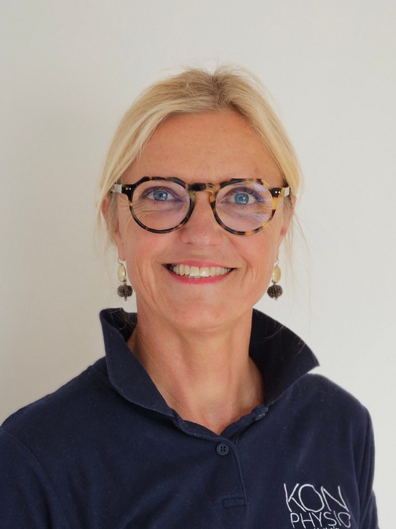 Physiotherapeutin Prien Kathrin Ortinger Naujeck Lächelnde Physiotherapeutin Kathrin Ortinger Naujeck mit blonden Haaren, Brille und blauer Kleidung vor einem hellen Hintergrund.