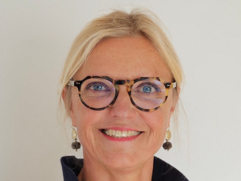 Physiotherapie Prien Kathrin Ortinger Naujeck Physiotherapeutin Kathrin Ortinger Naujeck mit blonden Haaren, Brille und blauem Polo mit KONPHYSIO Logo, lächelt freundlich in die Kamera.