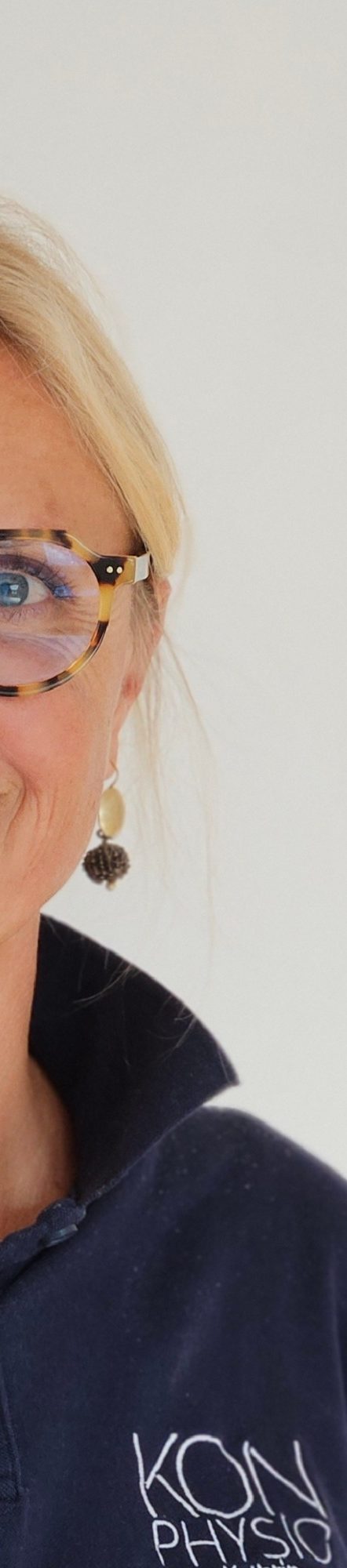 Physiotherapie Kathrin Ortinger Naujeck Prien am Chiemsee Physiotherapeutin Kathrin Ortinger Naujeck mit blonden Haaren, Brille und blauem Polo mit KONPHYSIO Logo, lächelt freundlich in die Kamera.