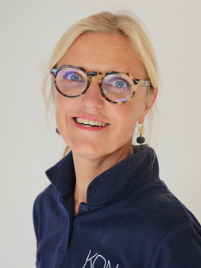 Physiotherapeutin Kathrin Ortinger Naujeck Prien am Chiemsee Physiotherapeutin Kathrin Ortinger Naujeck mit blonden Haaren, Brille und blauem Polo mit KONPHYSIO Logo, lächelt freundlich in die Kamera.