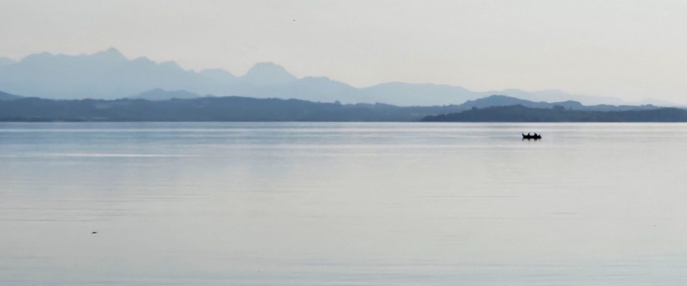 Physiotherapie Prien Kathrin Ortinger Naujeck Ruhiger Chiemsee mit sanften Bergen im Hintergrund und einem Boot auf der Wasseroberfläche.