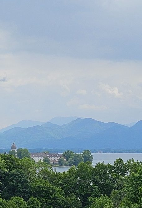 Physiotherapie am Chiemsee in Prien Kathrin Ortinger Naujeck Blick auf den Chiemsee mit Fraueninsel umgeben von Bäumen und Bergen unter einem heiter bewölkten Himmel.